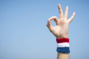 Hand of an athlete making OK sign with finger and thumb against blue sky wearing a red white and blue sport wristband